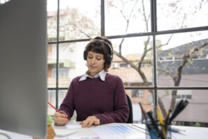 Woman wearing headphones working on media transcription at desk, taking notes while listening to audio on computer